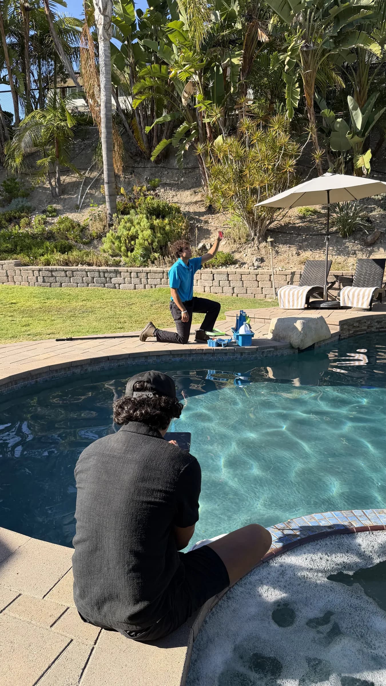 Technician in blue shirt kneels by backyard pool holding up a water test vial while another person sits at spa edge