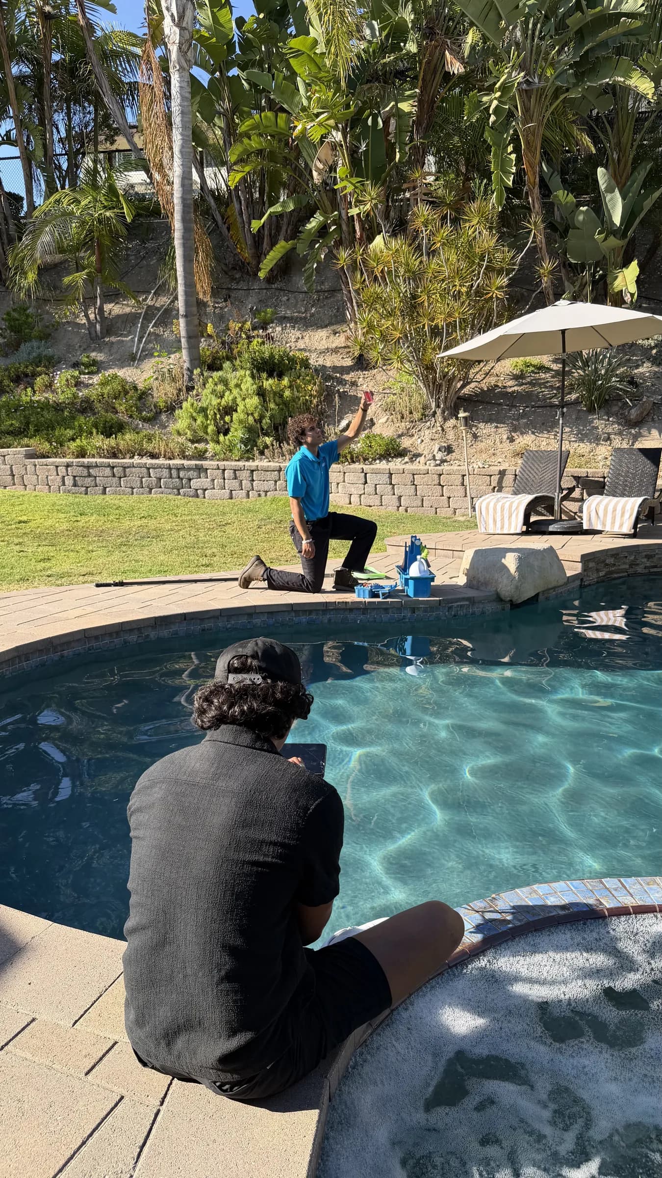 Technician in blue shirt kneels by backyard pool holding up a water test vial while another man sits at spa edge