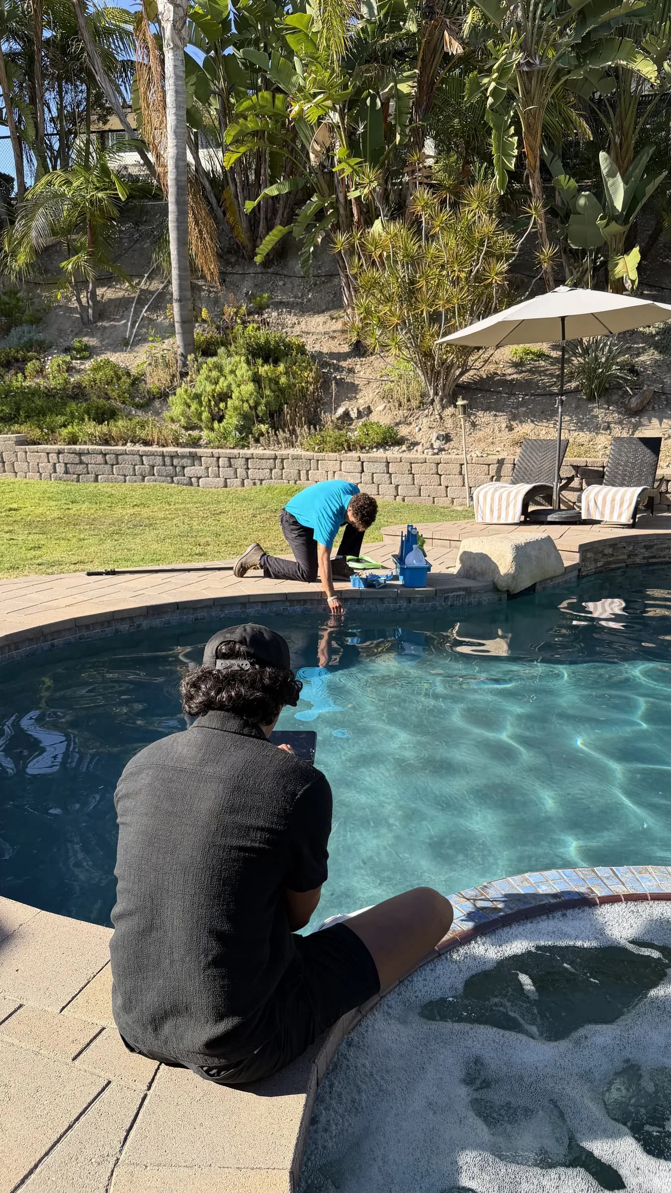 Technician kneeling at pool edge collecting water sample beside test kit while another person sits near bubbling spa