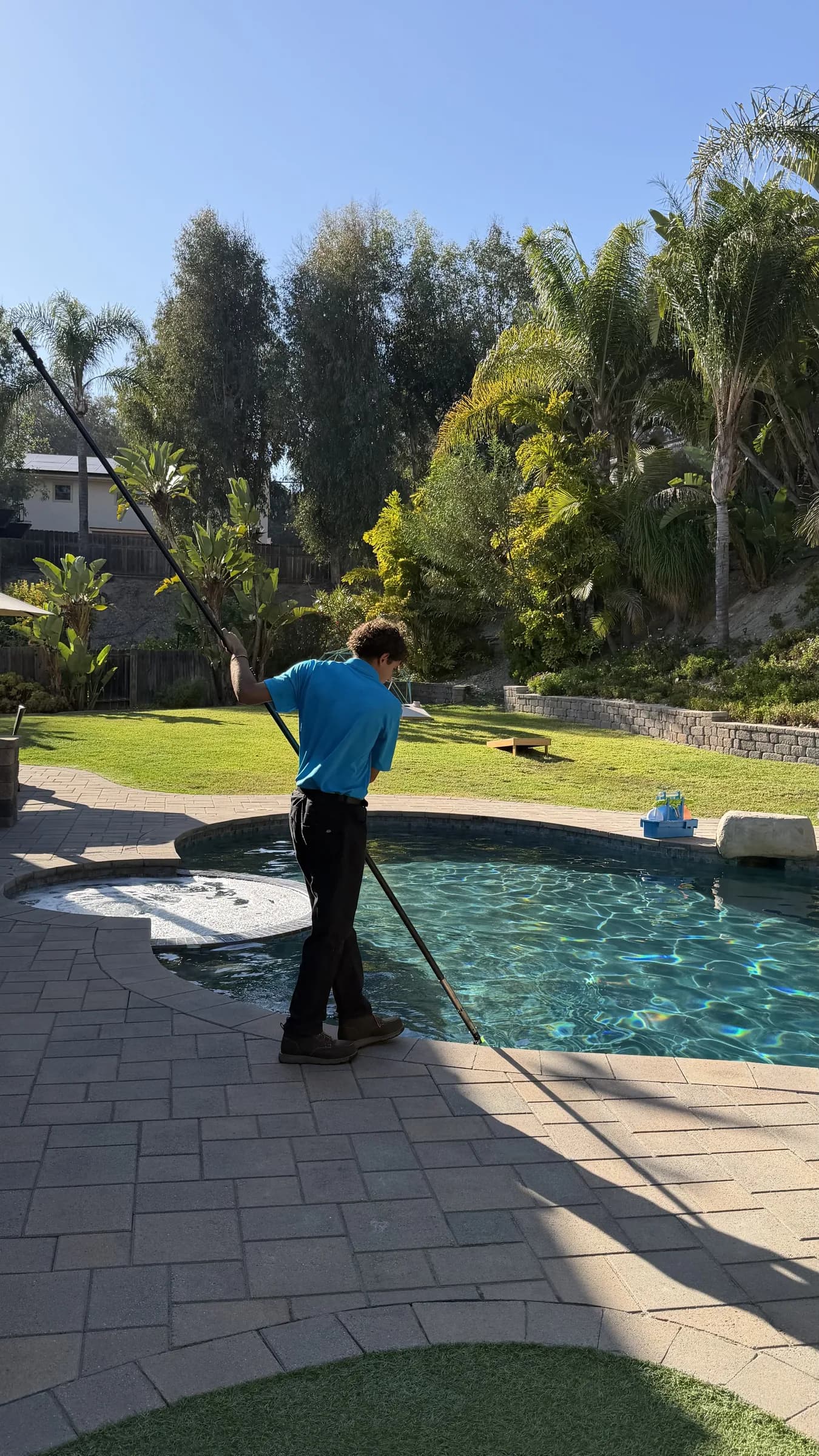 Pool technician in blue shirt using a long telescopic pole to clean a backyard pool beside a spa