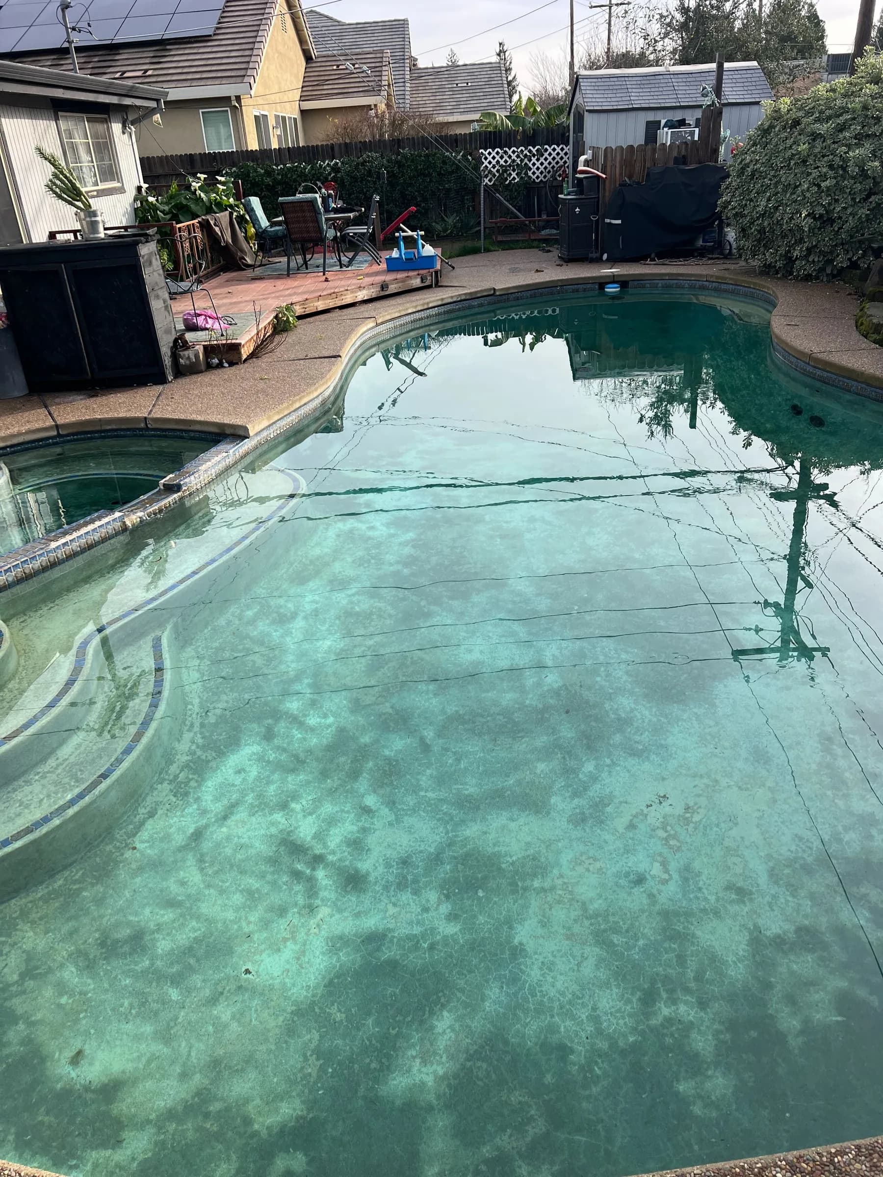 Cloudy green-tinted backyard pool with blotchy algae on the floor, attached spa, steps, and wooden deck with patio furniture