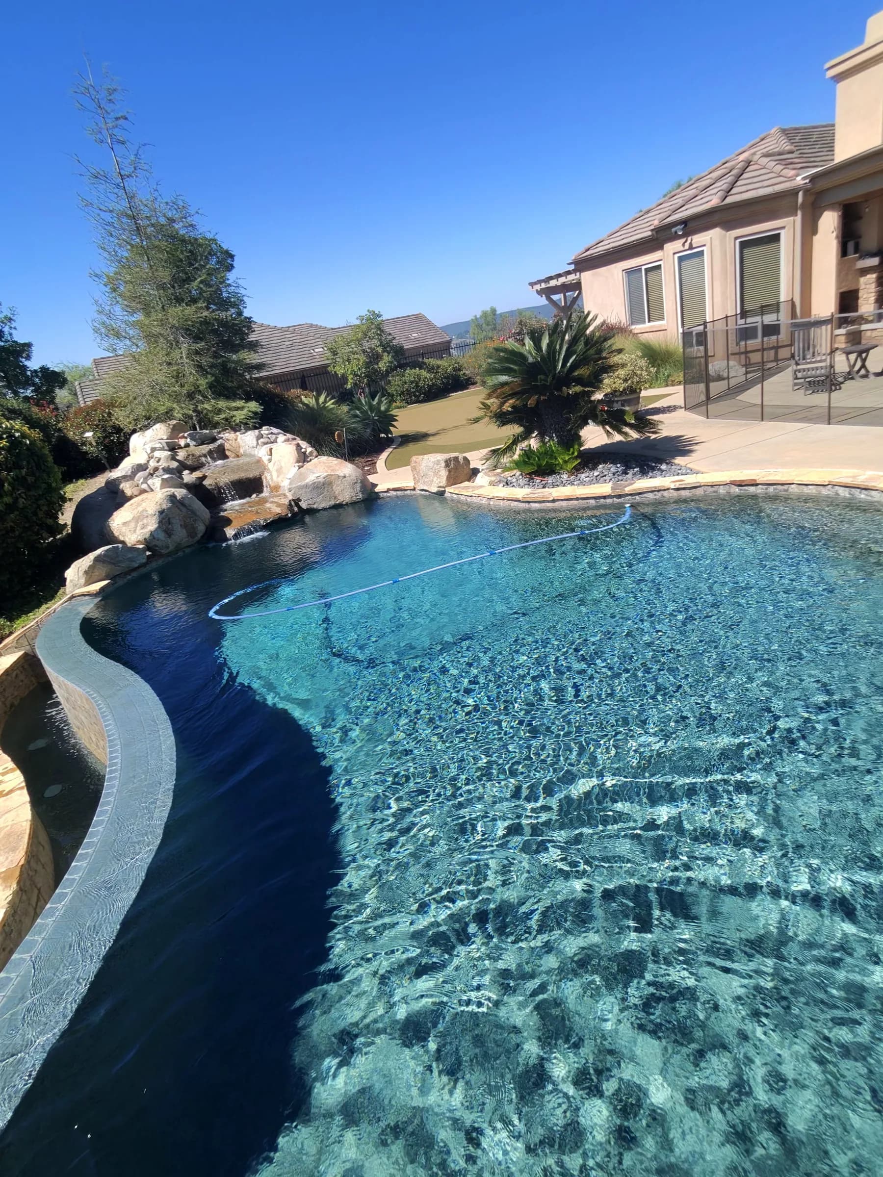 Clear blue backyard pool with rock waterfall, curved spa wall, floating cleaner hose, and stucco house under blue sky