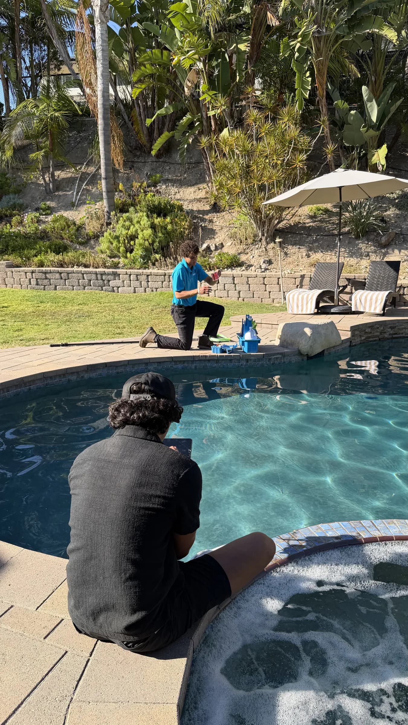 Technician in blue shirt kneels at pool edge testing water while another person sits by bubbling spa