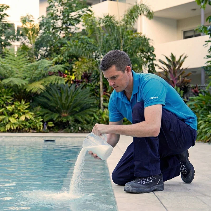 Technician in blue polo kneels at pool edge pouring white granular chemical from a plastic jug into water, tropical plants behind