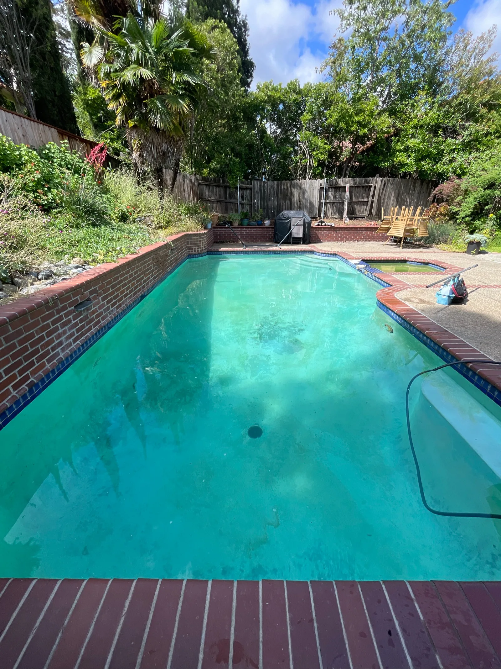 Rectangular backyard pool with cloudy teal water, brick coping, attached spa, vacuum hose, and wooden fence behind