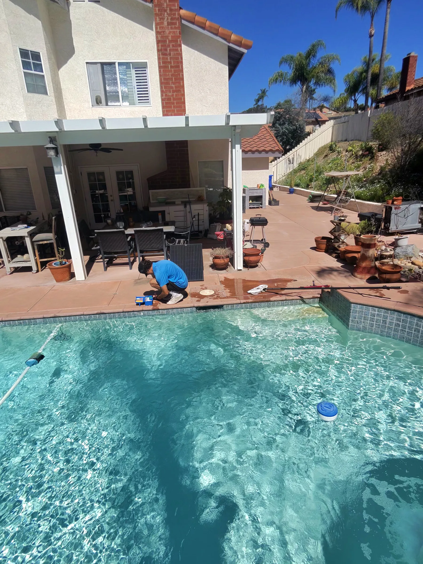 Technician in blue shirt crouches at pool edge with test kit beside clear turquoise backyard pool and covered patio