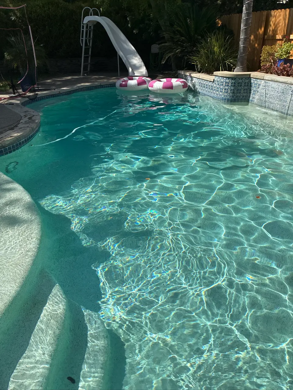 Sparkling turquoise pool with curved entry steps, a slide, floating rings, and a raised patterned tile wall, after pool service in Antelope, CA
