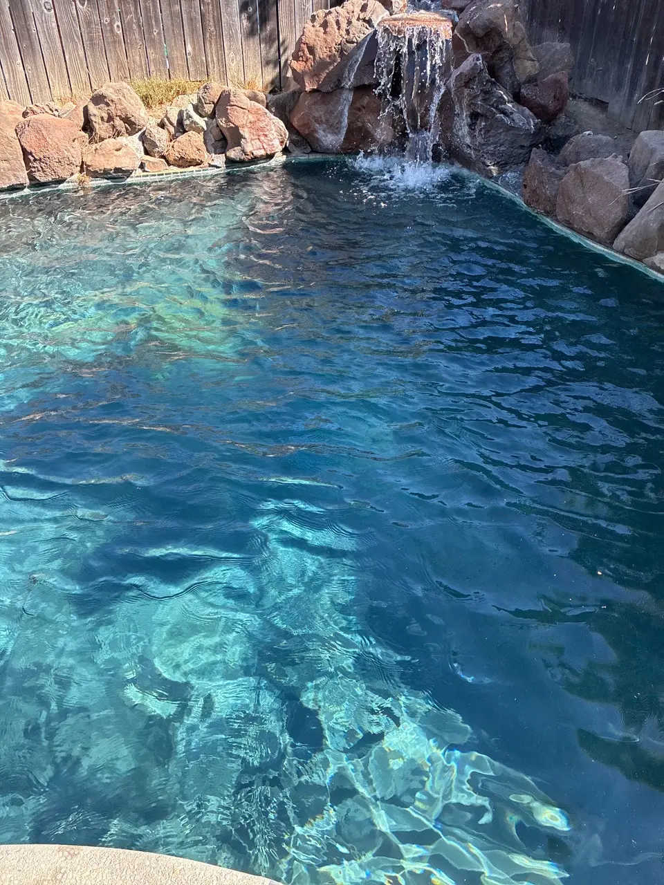 Sparkling blue pool water ripples beneath a flowing rock waterfall, bordered by natural stone and wooden fencing, after pool service in Antelope, CA