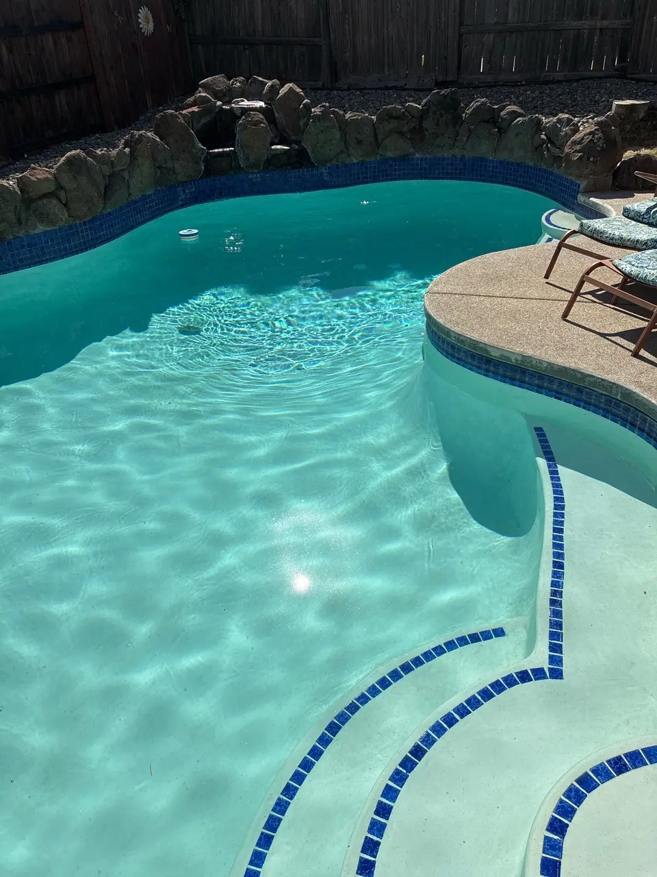 Sparkling turquoise water reveals curved blue-trimmed steps beneath a rock waterfall and rounded poolside seating area, after pool service in Antelope, CA