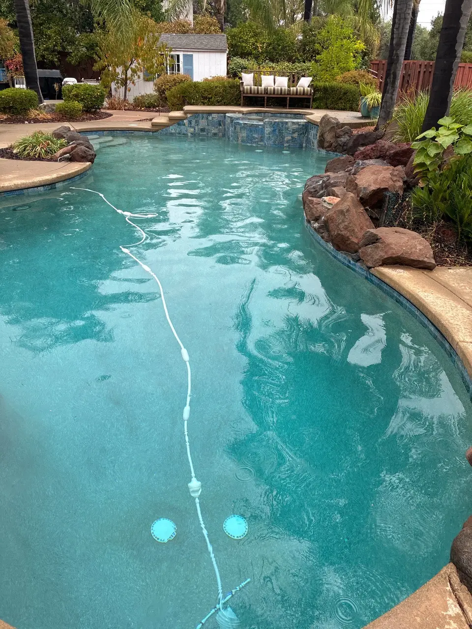 Clear turquoise water fills a freeform pool with a raised tiled spa, rock edging, and tropical landscaping, after pool service in Antelope, CA