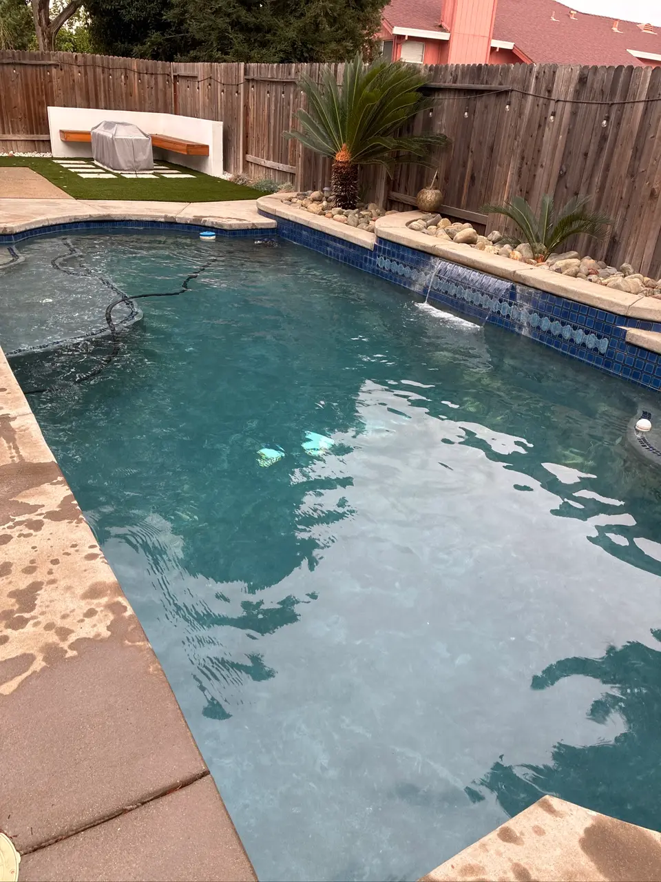 Clear turquoise pool water beneath a tiled waterfall wall, with stone coping, palms, and a submerged cleaner hose, after pool service in Antelope, CA
