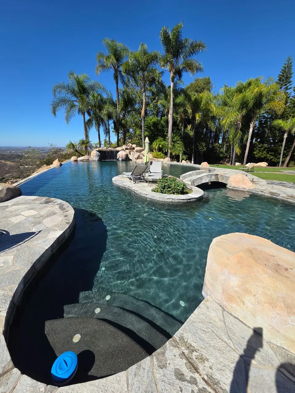 Sparkling teal water fills a dark-finished pool with stone bridge, island seating, rock waterfall, and surrounding palms, after pool service in Bonsall, CA