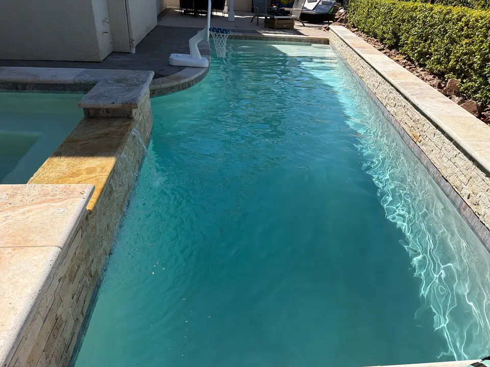 Clear turquoise pool water beside a raised spa, stone walls, and a poolside basketball hoop, after pool service in Carlsbad, CA