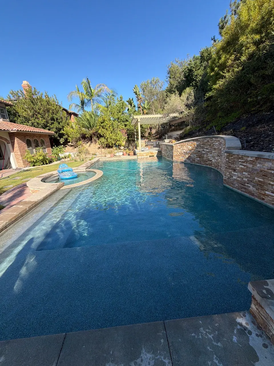 Sparkling blue pool with wide entry steps, an attached round spa, stone walls, and lush landscaping, after pool service in Carlsbad, CA