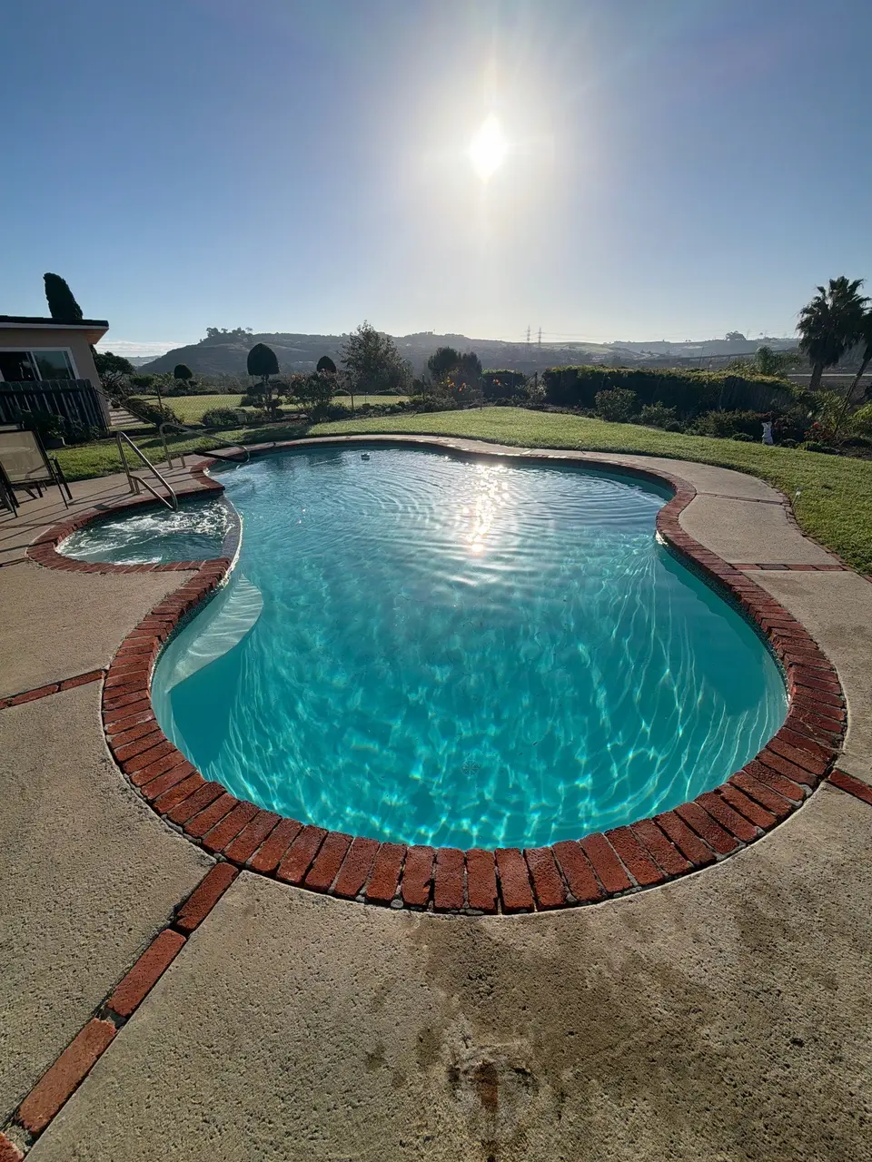 Sparkling turquoise water fills a curved pool with an attached bubbling spa, brick coping, and landscaped surroundings, after pool service in Carlsbad, CA
