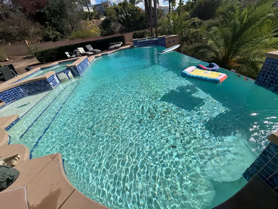 Sparkling turquoise pool with wide steps, blue tile accents, waterfall spillways, floating loungers, and surrounding palms, after pool service in Carlsbad, CA