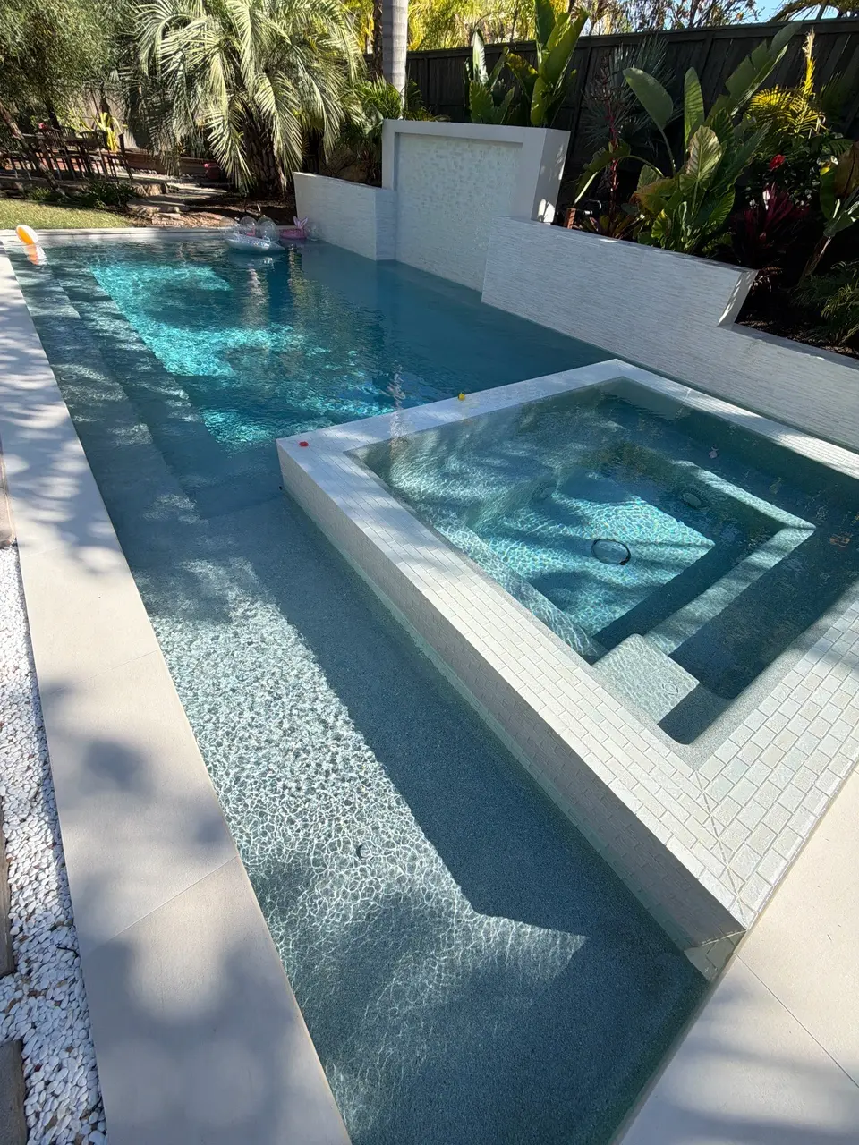 Sparkling blue water surrounds a raised tiled spa, with a white feature wall and tropical landscaping, after pool service in Chula Vista, CA