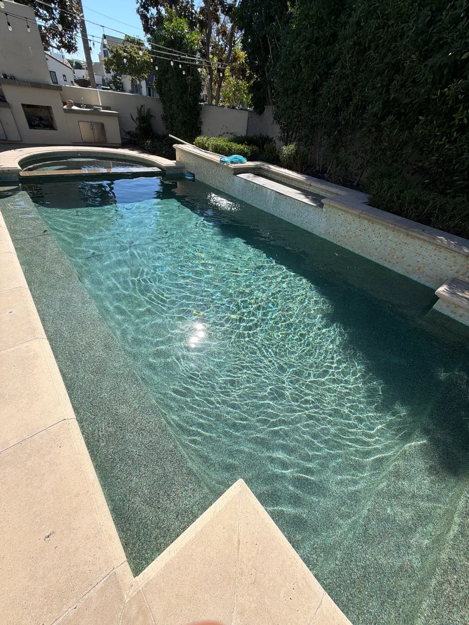 Sparkling turquoise water reveals a pebble finish, with a raised spa, tiled wall, and dense surrounding greenery, after pool service in Coronado, CA