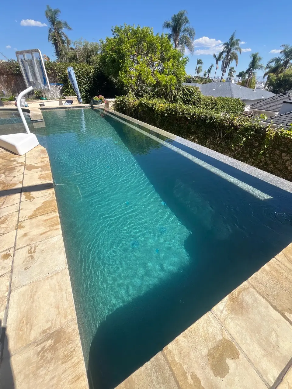 Pool with sparkling turquoise water, a long submerged bench, stone decking, and a basketball hoop amid tropical landscaping, after pool service in Coronado, CA