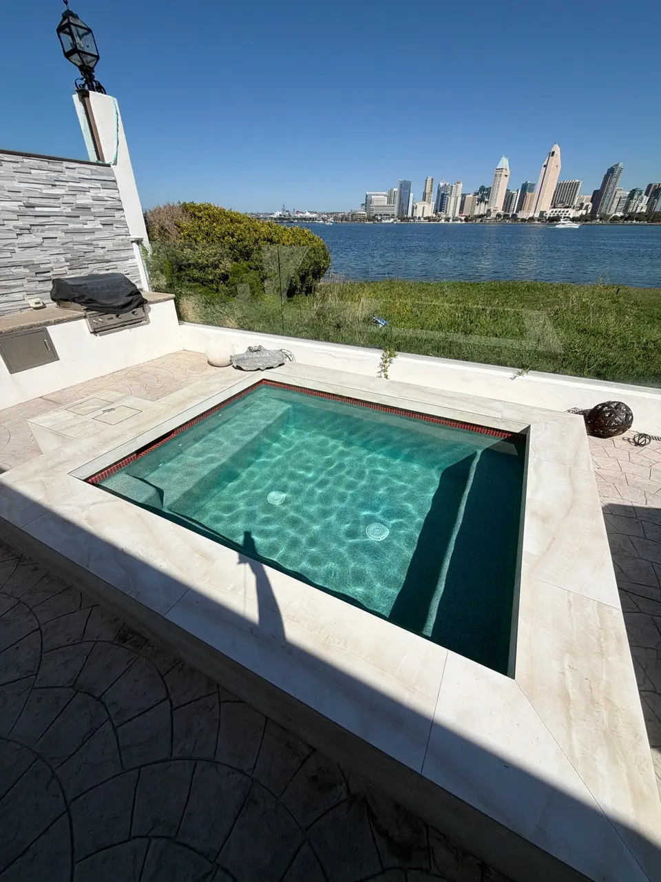 Square spa with sparkling turquoise water, submerged benches, and light stone coping overlooks a waterfront skyline, after pool service in Coronado, CA