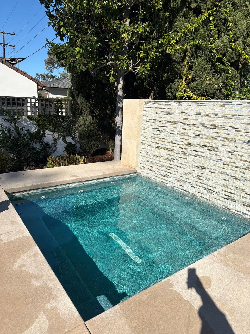 Rectangular spa with sparkling turquoise water, submerged seating, stone coping, and a mosaic tile wall beside trees, after pool service in Coronado, CA
