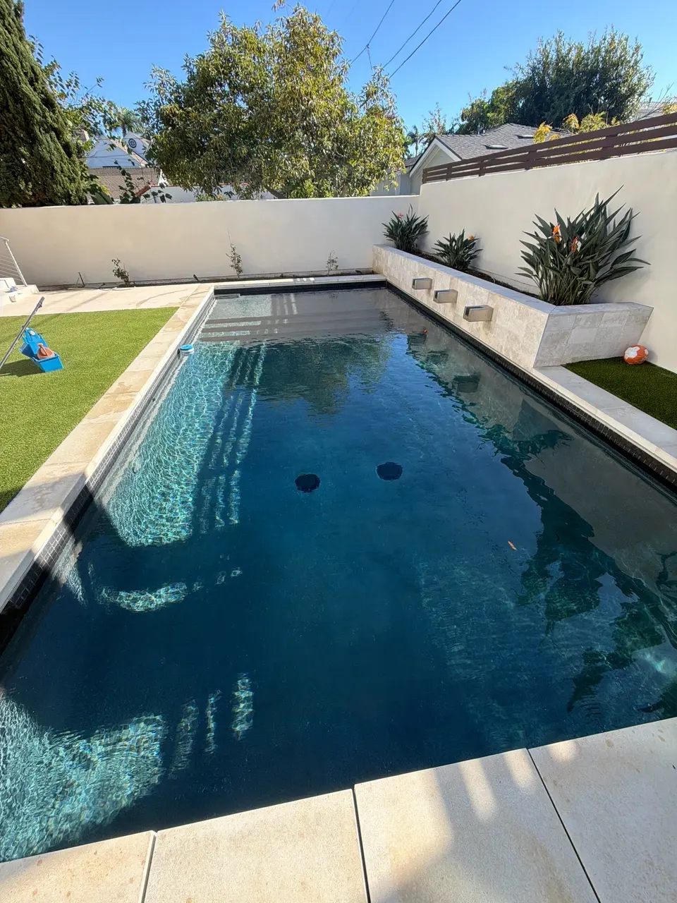 Clear blue water fills a rectangular pool with shallow steps, stone coping, and raised planters with spillways, after pool service in Coronado, CA