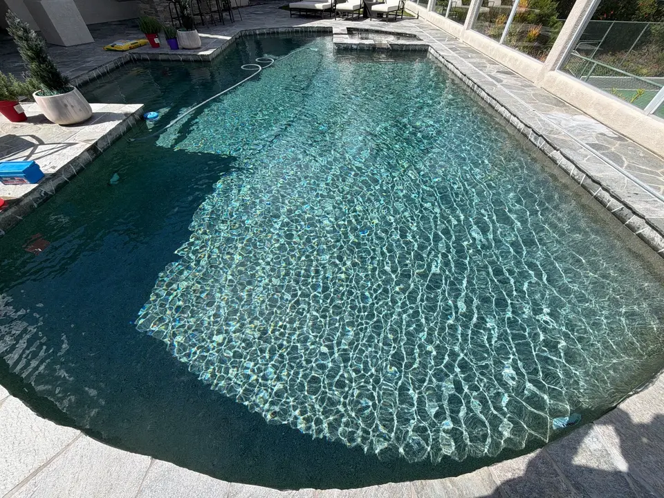 Sparkling blue water reveals a textured finish and entry steps in a screened pool with gray stone coping, after pool service in Del Mar, CA