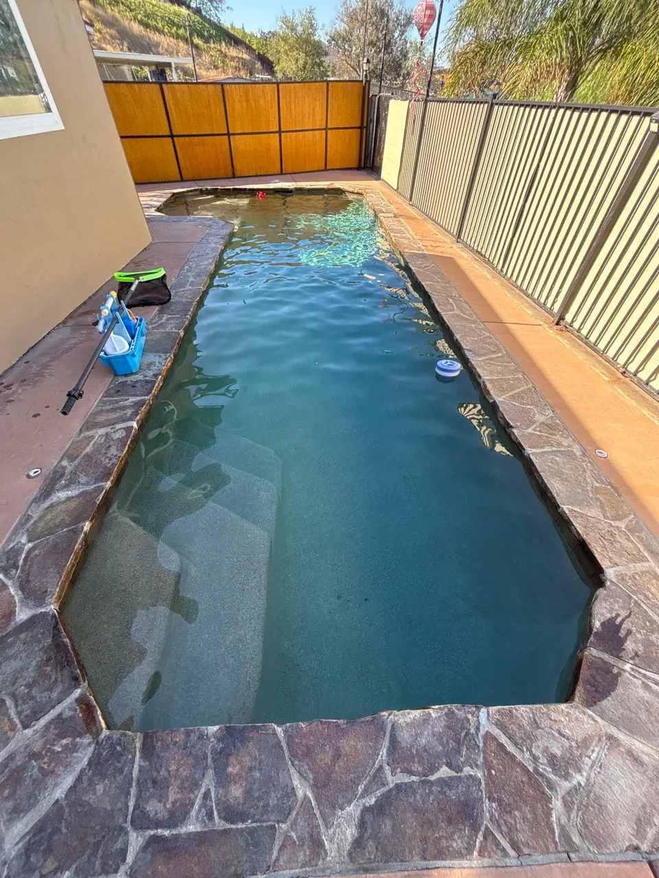 Clear blue water fills a narrow pool with submerged steps, stone coping, and surrounding safety fencing, after pool service in El Cajon, CA