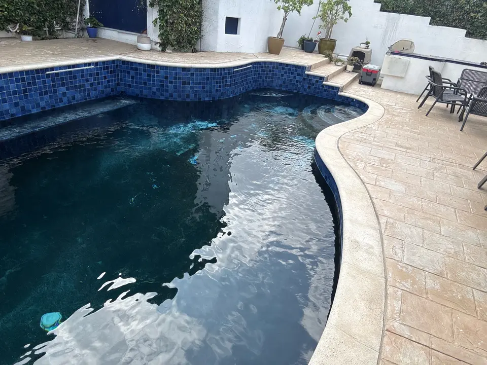 Clear dark teal water fills a curved pool with blue mosaic walls, entry steps, and light stone coping, after pool service in El Segundo, CA