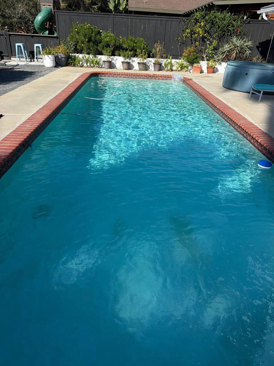 Sparkling turquoise water fills a rectangular pool with red brick coping, a water spout, and surrounding potted plants, after pool service in Encinitas, CA