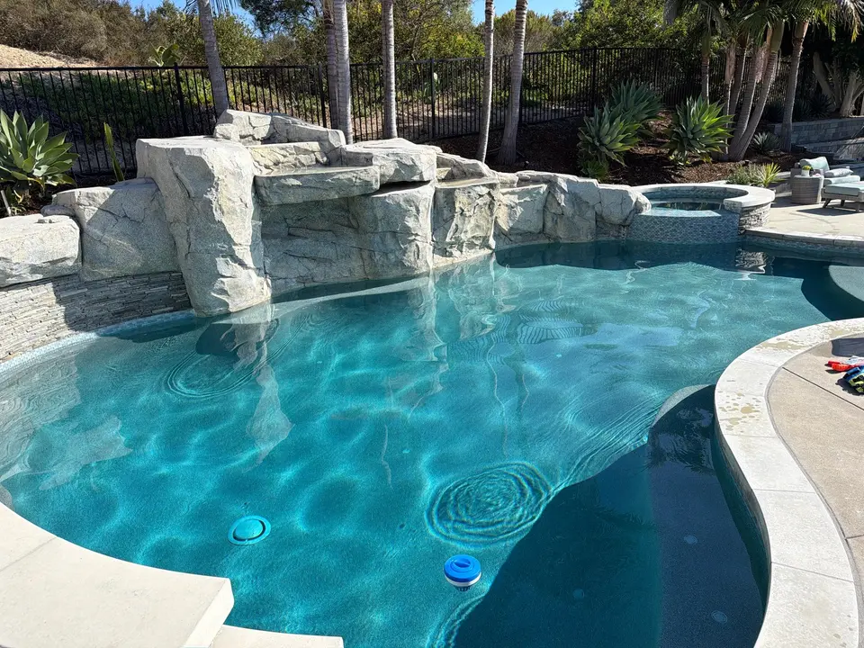 Sparkling turquoise pool with a raised tiled spa, large rock waterfall structure, and surrounding palms, after pool service in Encinitas, CA