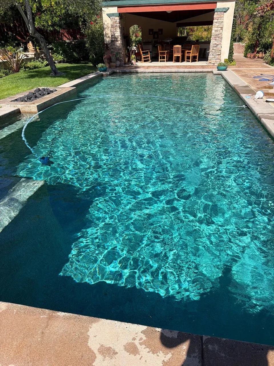 Sparkling turquoise pool water reveals submerged steps, with stone coping, landscaping, and a covered outdoor bar beyond, after pool service in Encinitas, CA