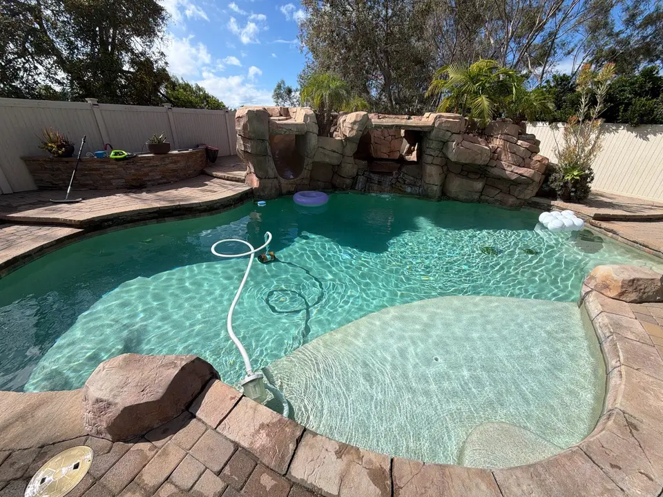Sparkling turquoise pool with a shallow sun shelf, rock waterfall grotto, stone coping, and tropical landscaping, after pool service in Encinitas, CA