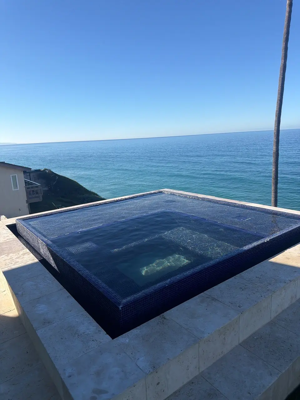 Clear water fills a raised, dark-tiled square spa with submerged seating, stone surround, and an ocean backdrop, after pool service in Encinitas, CA