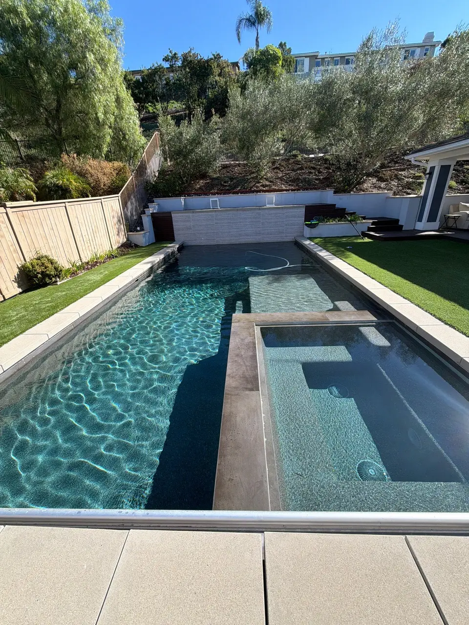 Sparkling blue water fills a rectangular pool with an integrated spa, shallow shelf, and landscaped hillside surroundings, after pool service in Encinitas, CA