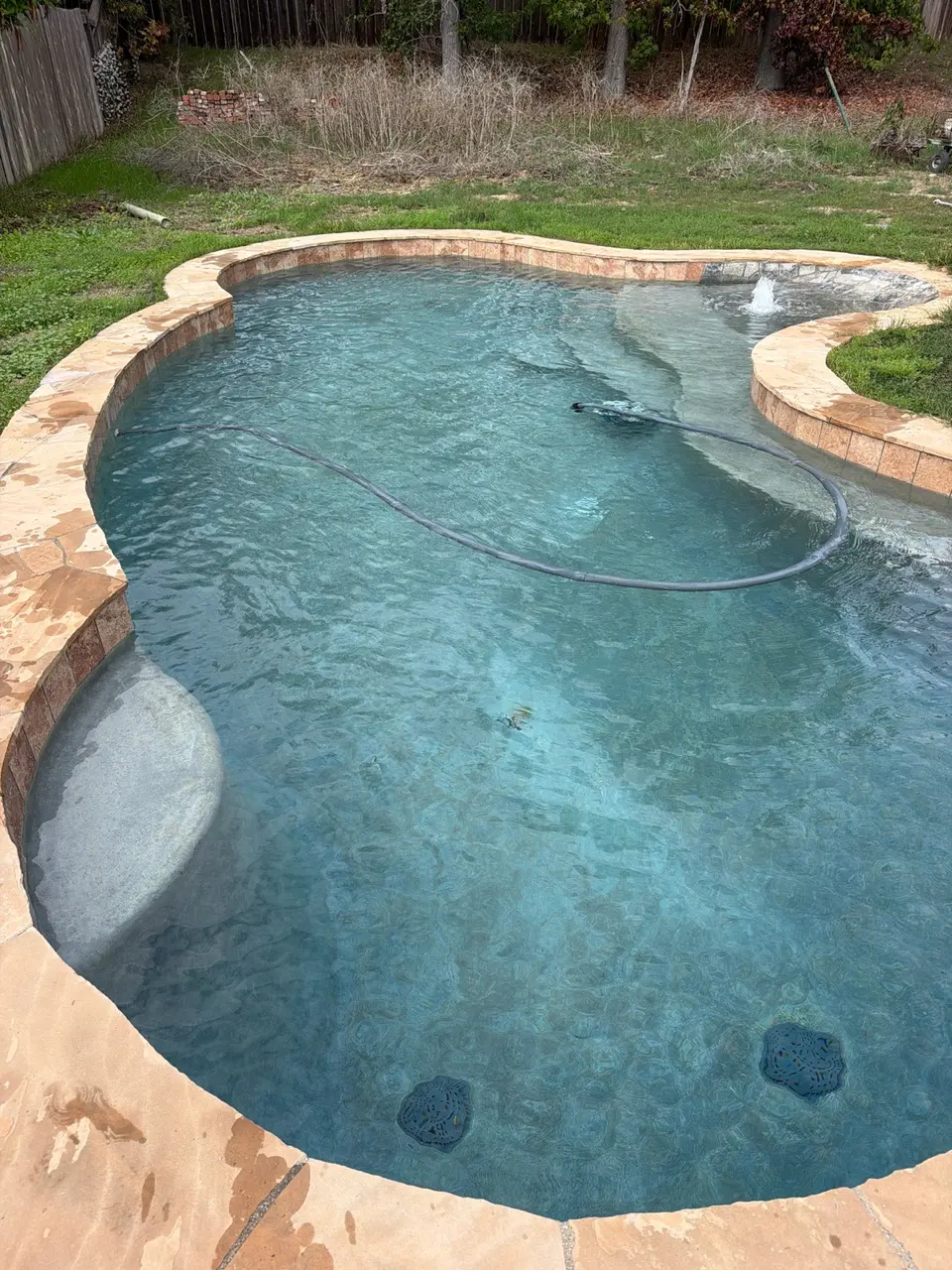 Clear blue water fills a freeform pool with stone coping, a submerged bench, and a bubbling sun shelf, after pool service in Encinitas, CA