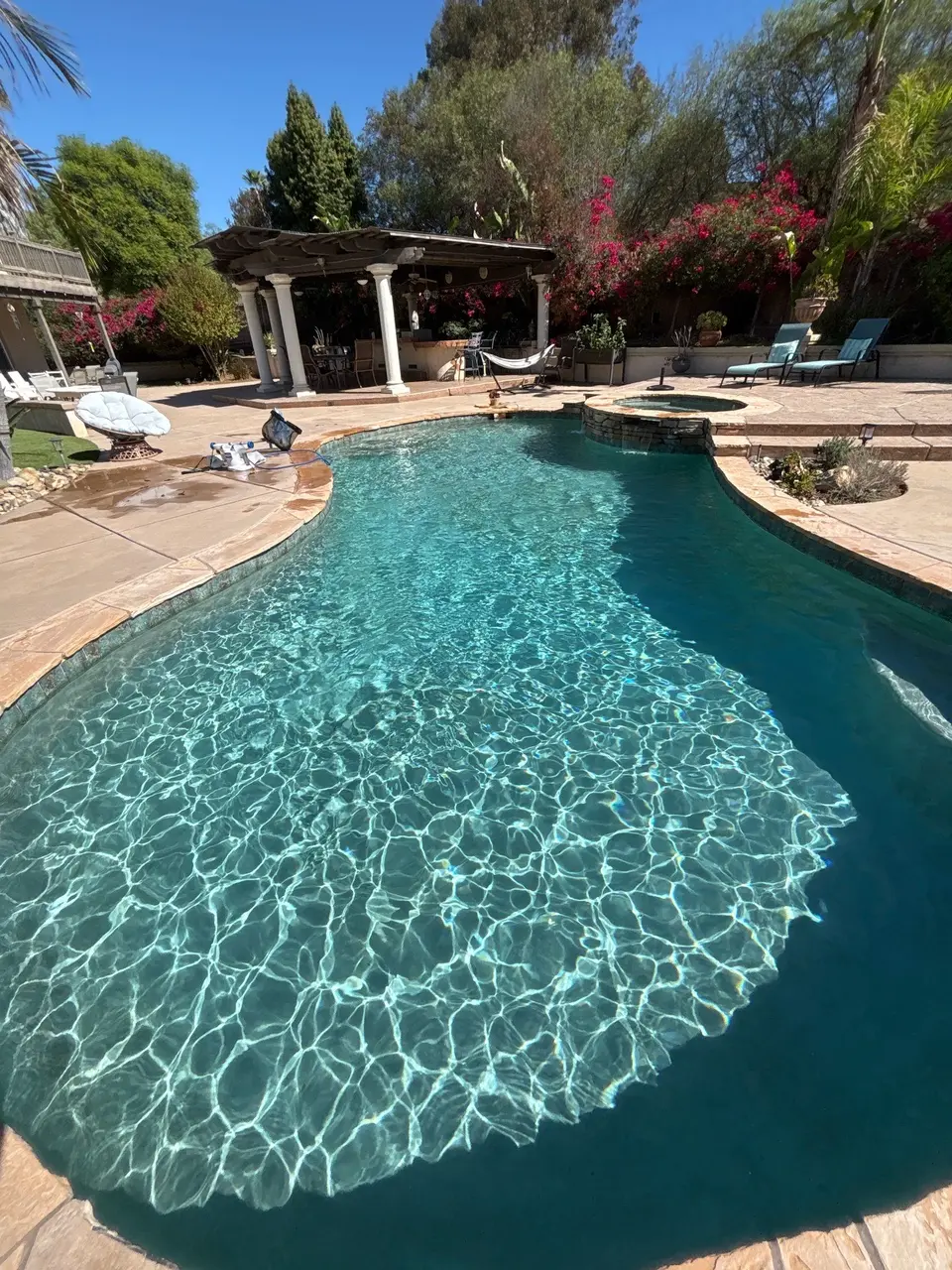 Sparkling turquoise water fills a freeform pool with a raised circular spa, stone coping, and flowering landscaping, after pool service in Escondido, CA