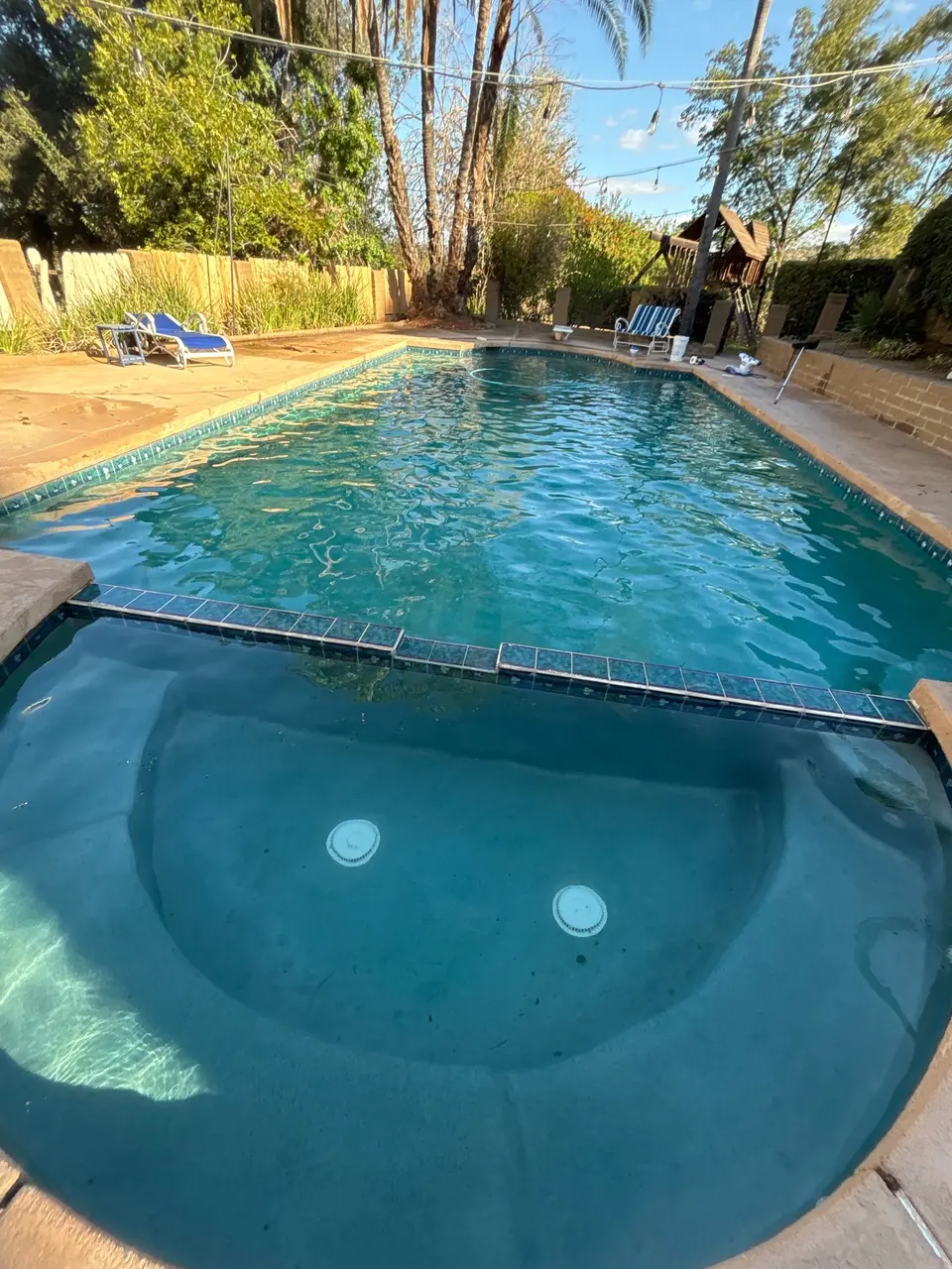 Clear blue water fills a backyard pool and rounded spa bordered by blue tile, stone decking, and mature landscaping, after pool service in Escondido, CA