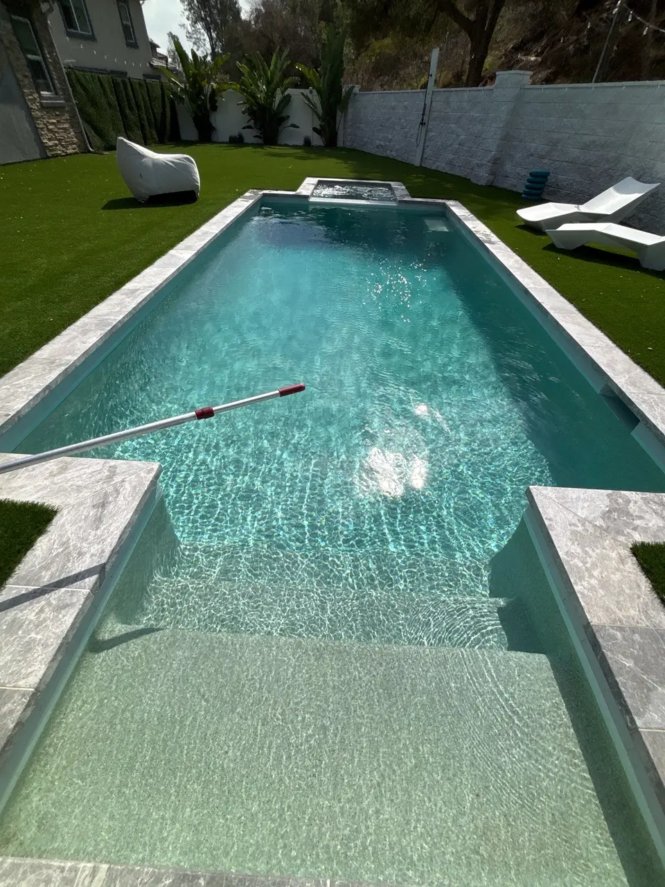 Sparkling turquoise water fills a rectangular pool with wide entry steps, light stone coping, and a raised spa, after pool service in Escondido, CA