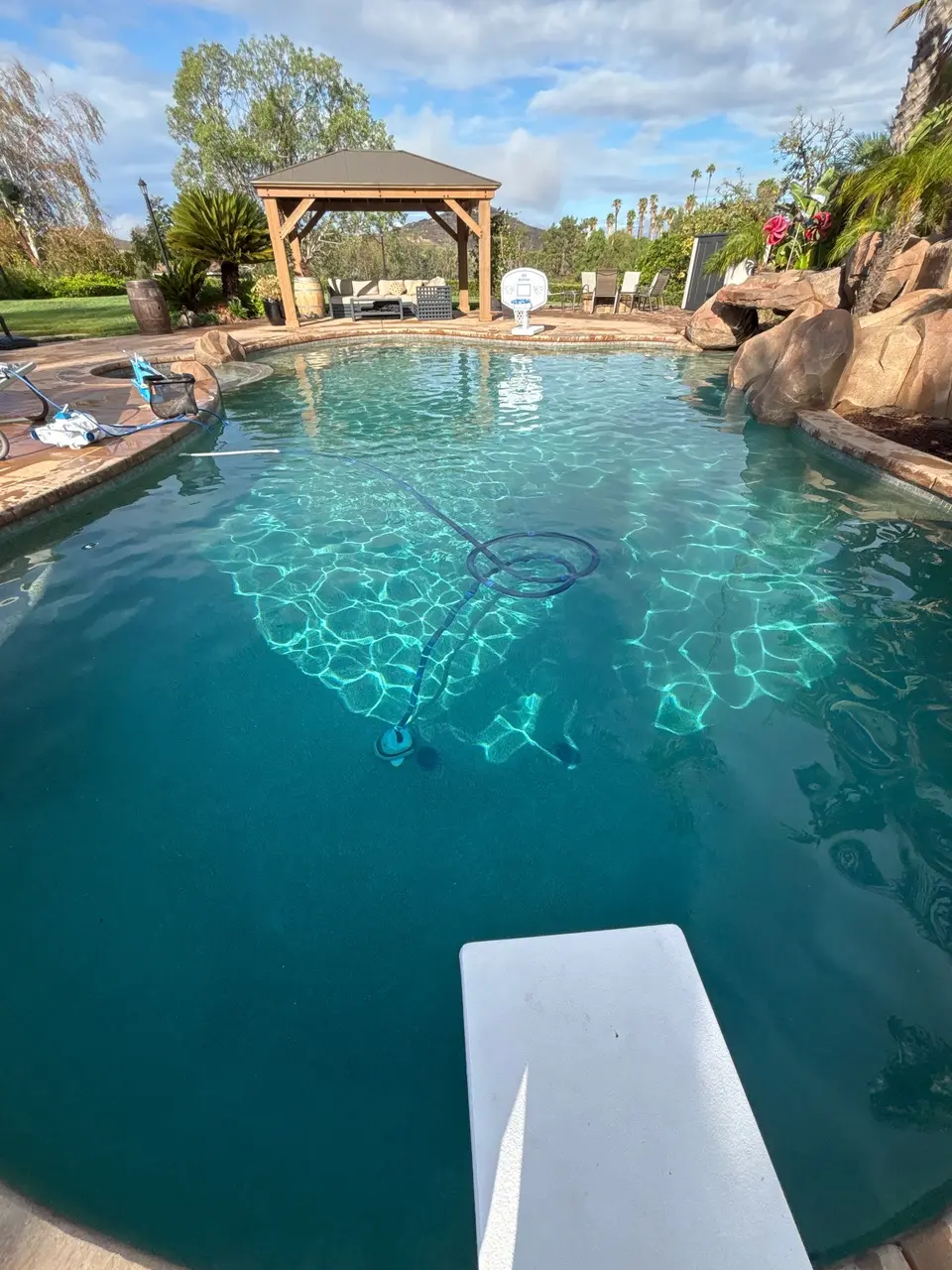 Sparkling turquoise pool with a diving board, rock grotto, cleaning hose, gazebo, and tropical landscaping, after pool service in Escondido, CA