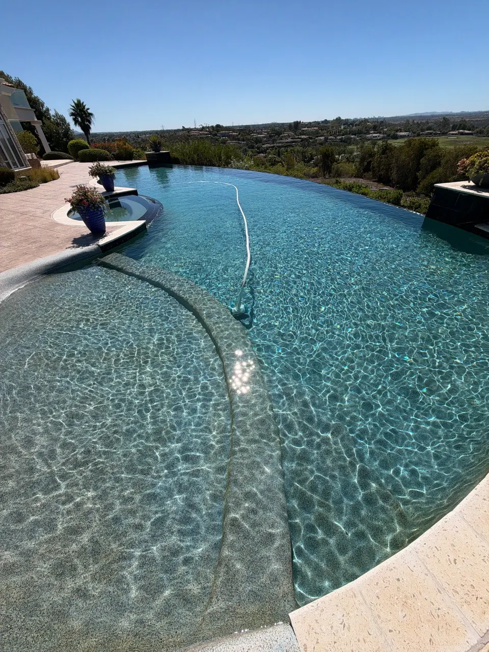 Sparkling turquoise pool with an infinity edge, curved shallow entry, pebble finish, and decorative planters overlooking greenery, after pool service in Fairbanks Ranch, CA