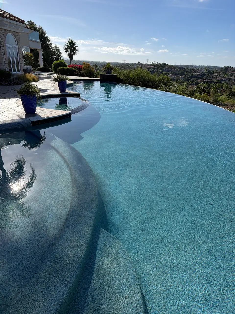 Sparkling blue infinity pool with curved submerged steps, an integrated spa, decorative planters, and hillside views, after pool service in Fairbanks Ranch, CA