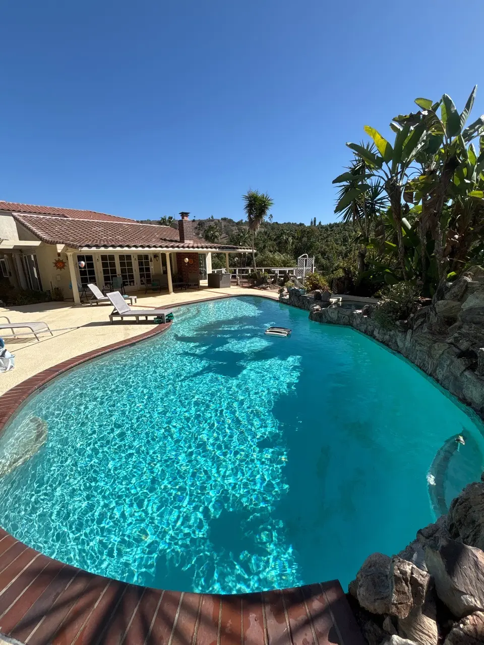 Freeform pool with sparkling turquoise water, brick coping, a raised rock-bordered spa, and tropical landscaping, after pool service in Fallbrook, CA