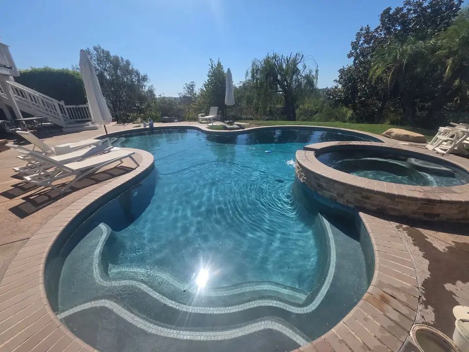 Sparkling blue water fills a freeform pool with curved tiled steps, a raised stone spa, and surrounding landscaping, after pool service in Fallbrook, CA