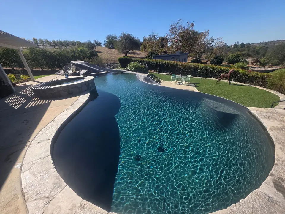 Sparkling blue water fills a freeform pool with a raised stone spa, rock waterfall feature, and landscaped surroundings, after pool service in Fallbrook, CA