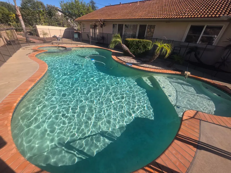 Sparkling turquoise water fills a freeform pool with an attached spa, broad steps, brick coping, and palm landscaping, after pool service in Fallbrook, CA