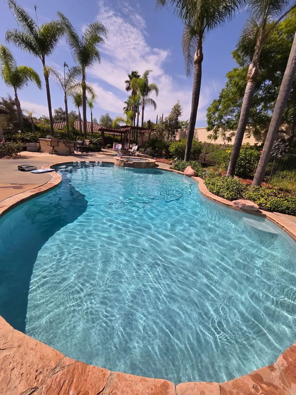 Sparkling blue water fills a freeform pool with a raised spa, stone coping, and surrounding palms, after pool service in Fallbrook, CA