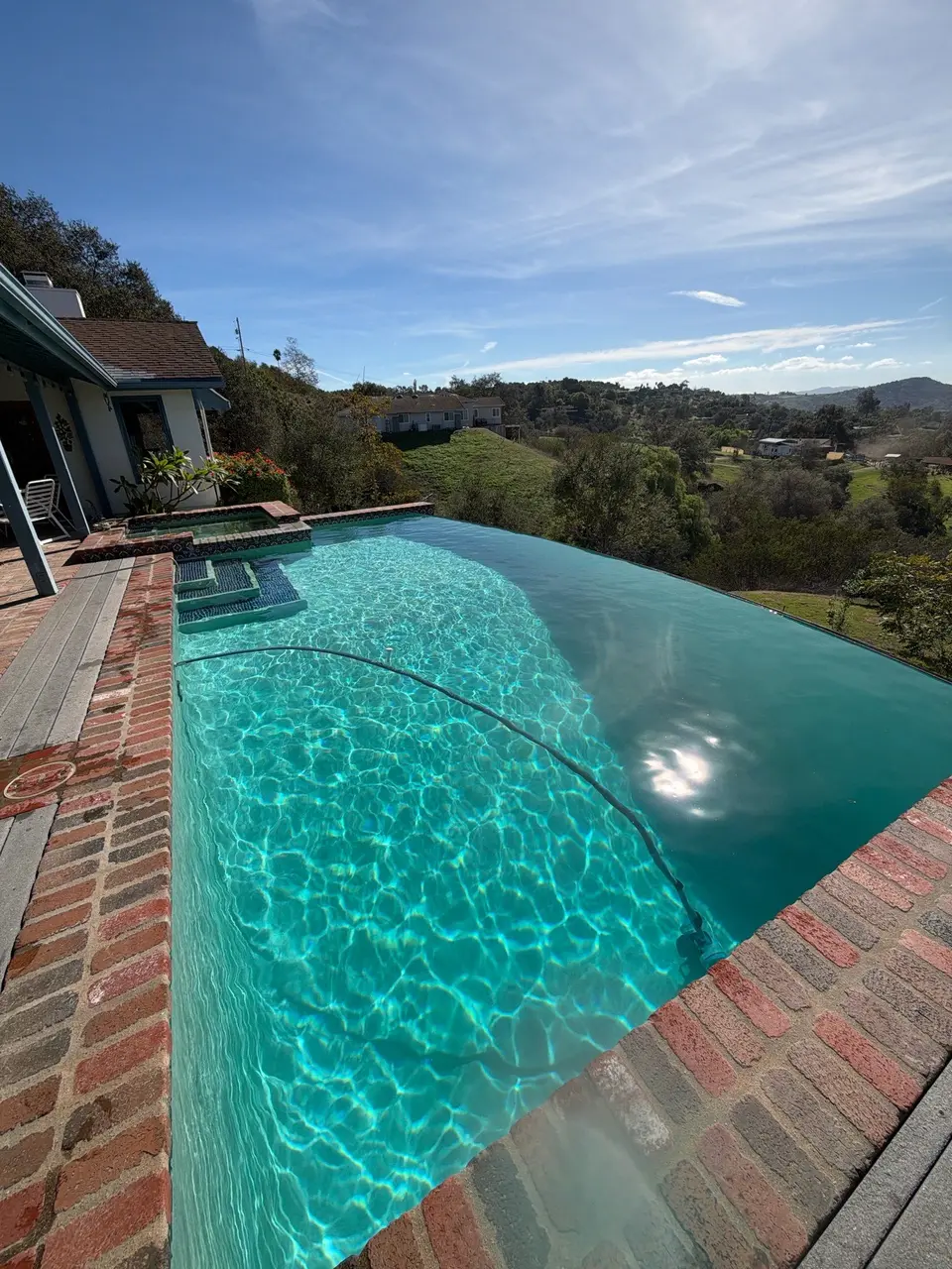 Sparkling turquoise water fills an infinity-edge pool with brick coping, tiled steps, and a raised spa overlooking hills, after pool service in Fallbrook, CA