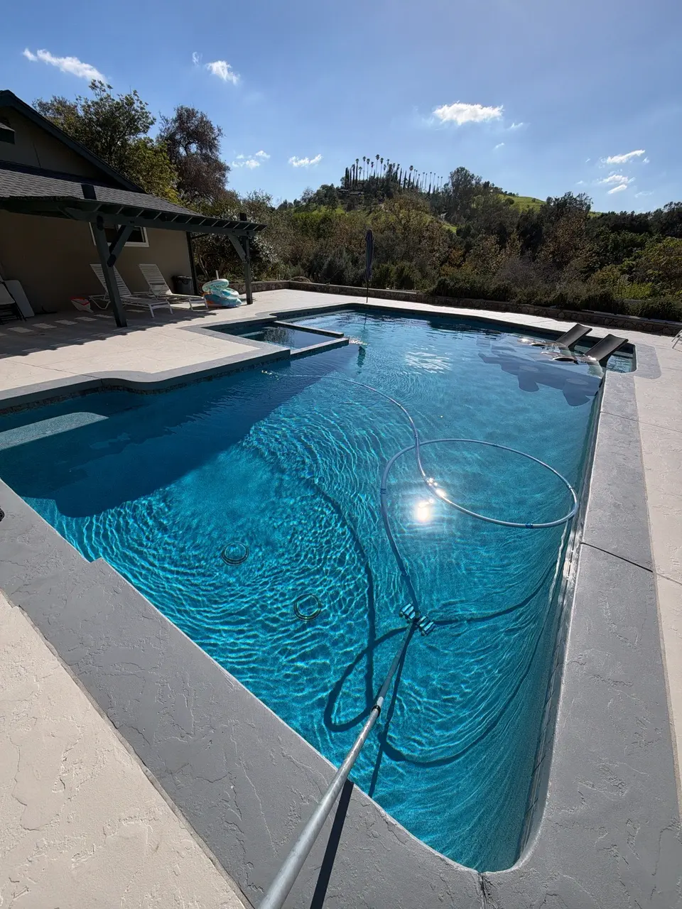 Sparkling blue pool with an attached spa, entry steps, submerged loungers, and landscaped hillside surroundings, after pool service in Fallbrook, CA