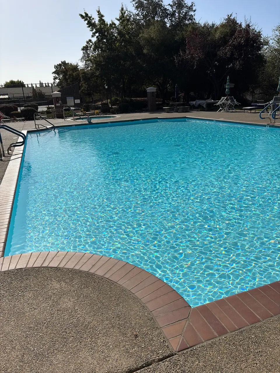 Sparkling turquoise pool water bordered by brick coping, metal handrails, lounge chairs, and trees, after pool service in Folsom, CA